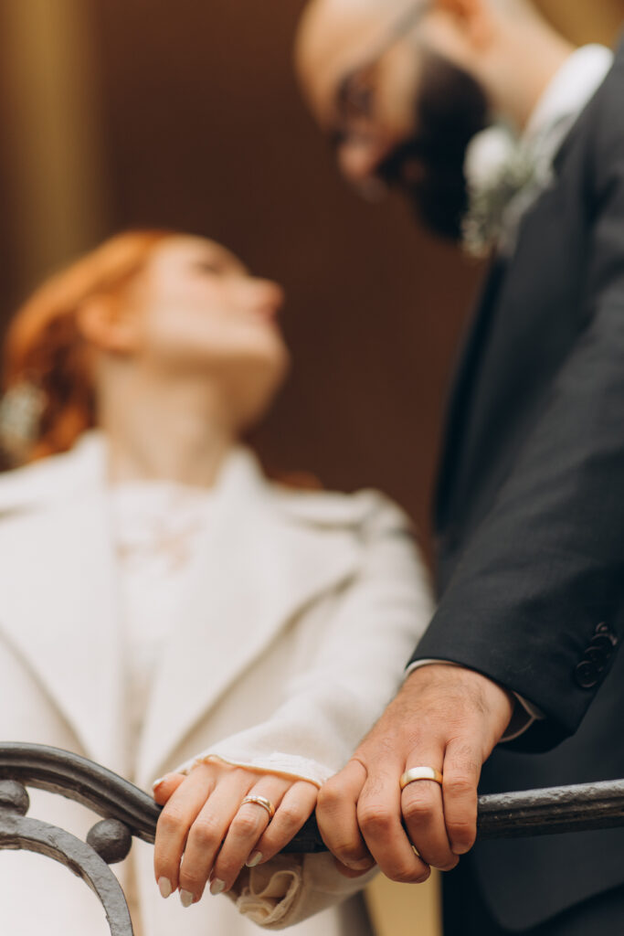 Detailaufnahme von Händen mit Eheringen, im Hintergrund unscharf ein verliebtes Hochzeitspaar, emotionale Hochzeitsfotografie mit Fokus auf die kleinen Momente.